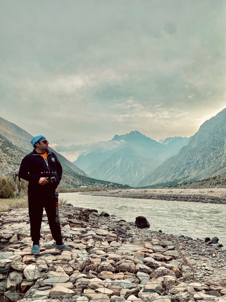 Dr. Mohit standing beside a river in a Himalayan valley, mountains rising behind him.